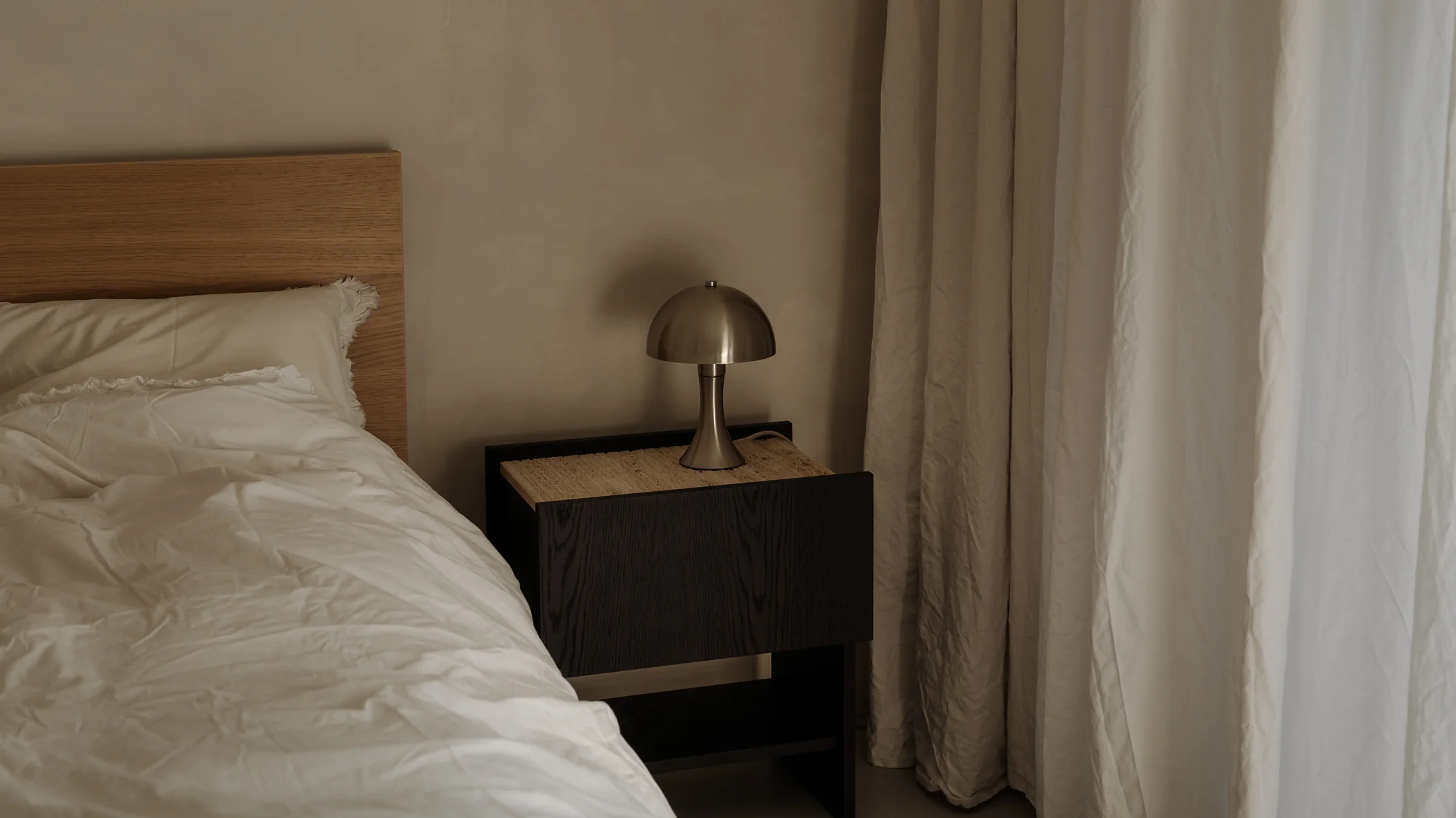 Chambre avec lit défait, lampe en dôme sur la table de chevet et rideaux clairs