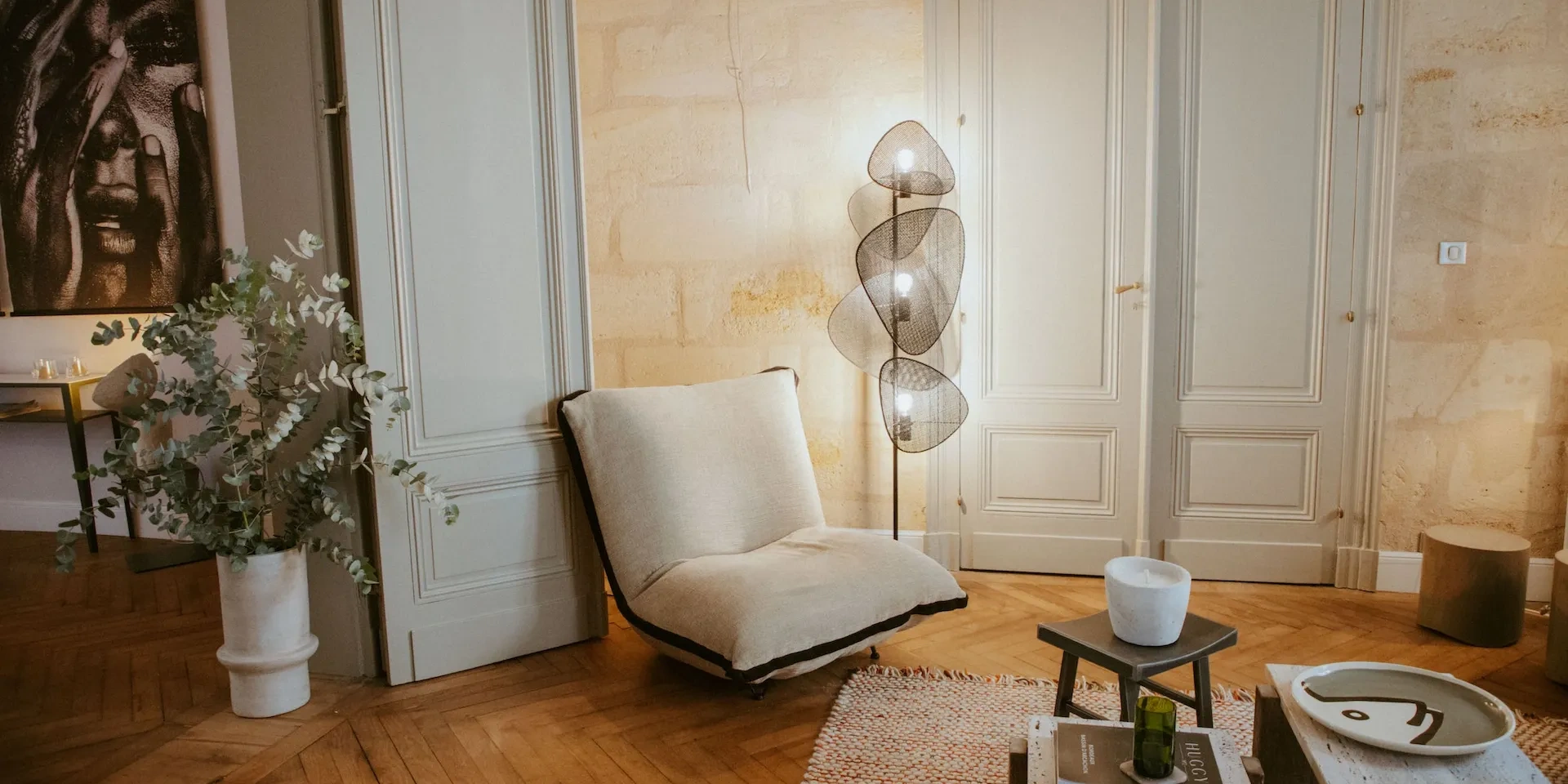 Un coin du salon de Maison Mandel : fauteuil blanc, lampadaire et portes anciennes sur un mur en pierre