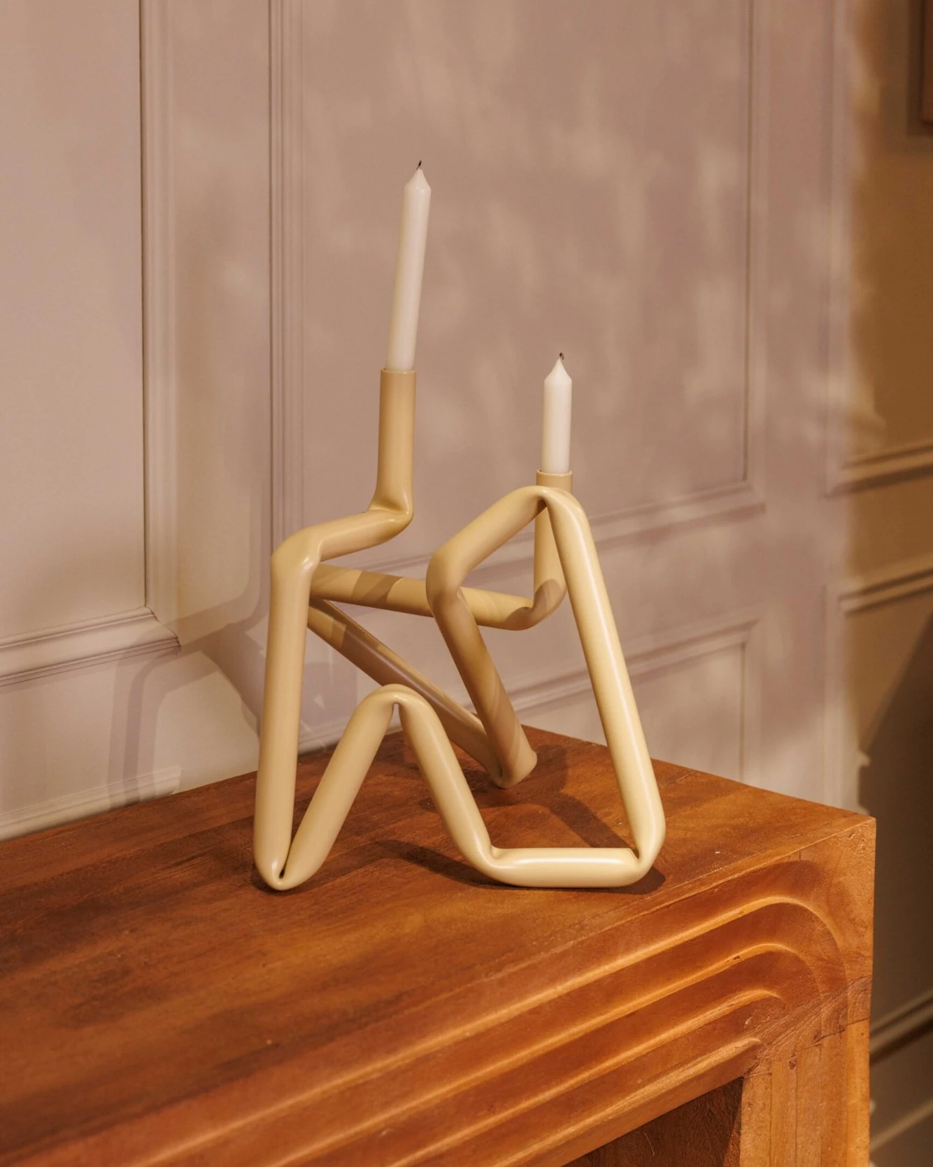 Un bougeoir en tube beige avec deux bougies, posé sur une console en bois devant un mur à moulures
